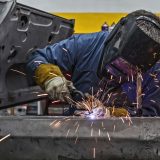 Man with salt and pepper gray hair wears face shield while welding in an automobile manufacturing plant which has undergone facility retrofit with Ionic Blue LLC