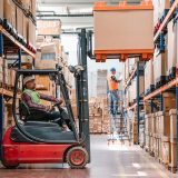 A worker wearing a neon yellow vest and hard hat uses a fork lift to move a large box within a distribution center