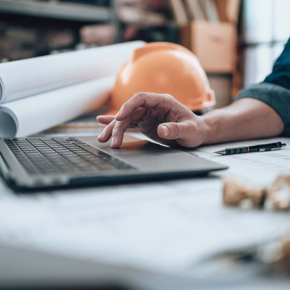 An individuals hands using the mouse pad of a laptop with architectural plans for a facility retrofit with IonicBlue LLC and an orange hard hat in the background