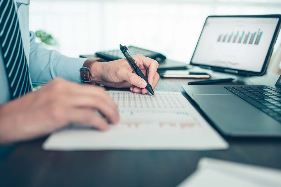 Close up on a man's hands holding a pen marking off items on a printed excel sheet reviewing infrastructure financing with IonicBlue LLC